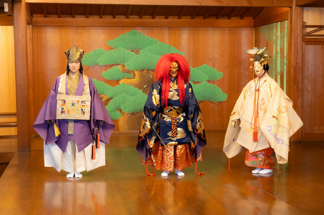 Three Japanese Noh performers standing in a row on a traditional wooden stage with a painted pine tree backdrop. The figure on the left is a man without a mask, wearing a purple robe and a tall gold headpiece. The central figure wears a fierce gold mask, a long vibrant red wig, and a dark blue robe with gold cloud patterns. The figure on the right portrays a female character with a white mask, wearing an ornate cream-colored kimono with floral patterns and a decorative metallic headdress.