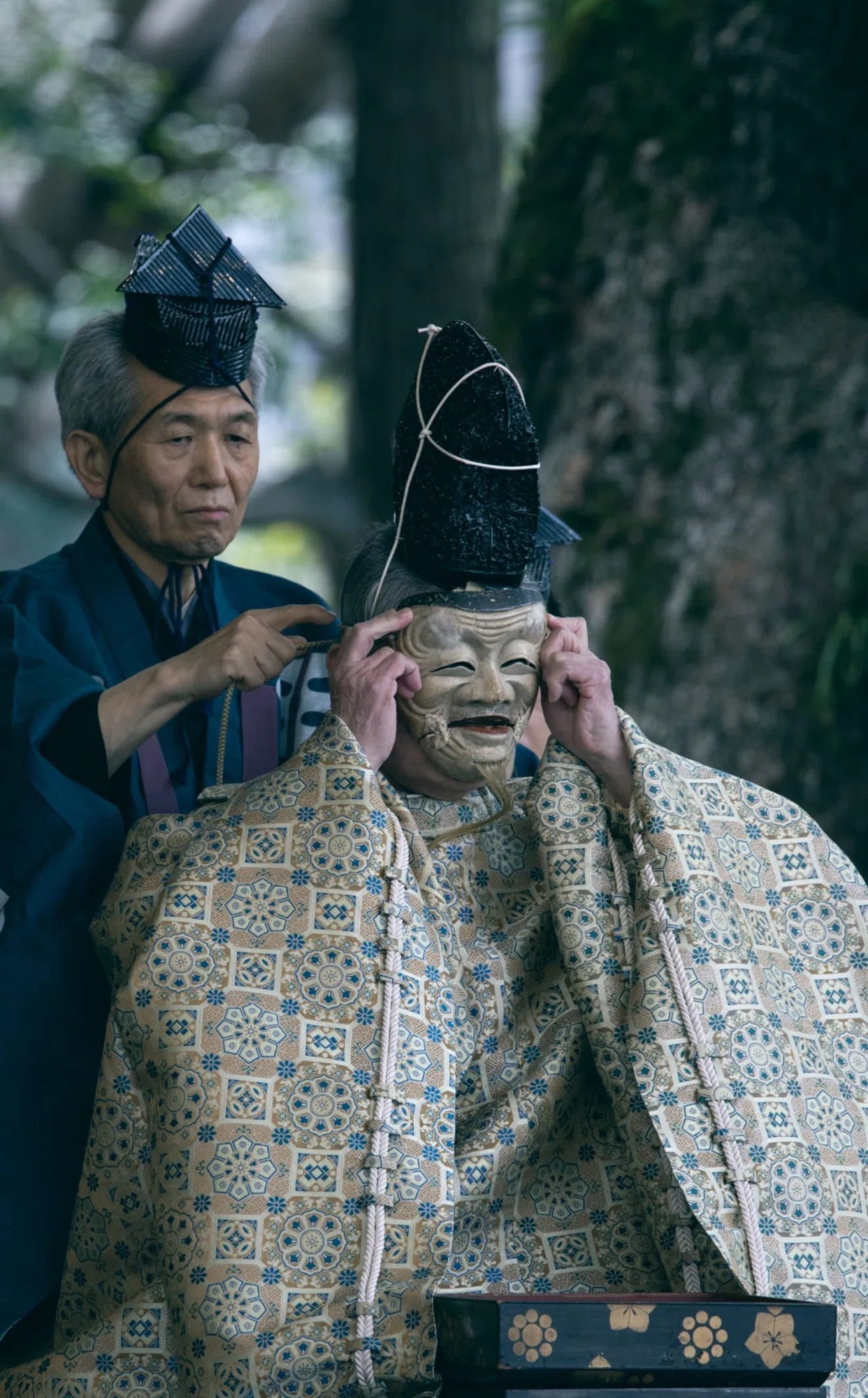 An intimate moment behind the scenes of a Noh performance. A seated performer, wearing a tall black hat (eboshi) and an intricately patterned cream robe, has an "Okina" (old man) mask carefully adjusted by an assistant in a blue garment. The setting appears to be outdoors with trees in the blurred background, capturing a quiet sense of preparation.