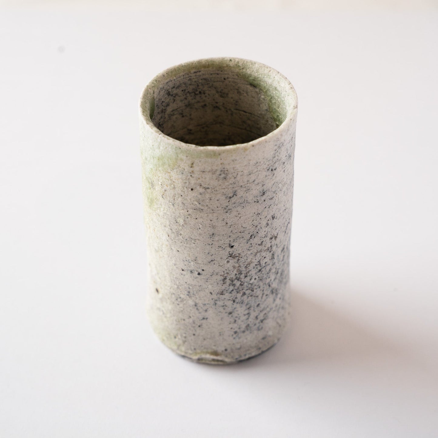 High-angle perspective of the white, granular side of Yui Tsujimura’s cylindrical YUNOMI cup, showing delicate dark mineral specks across the ceramic body.
