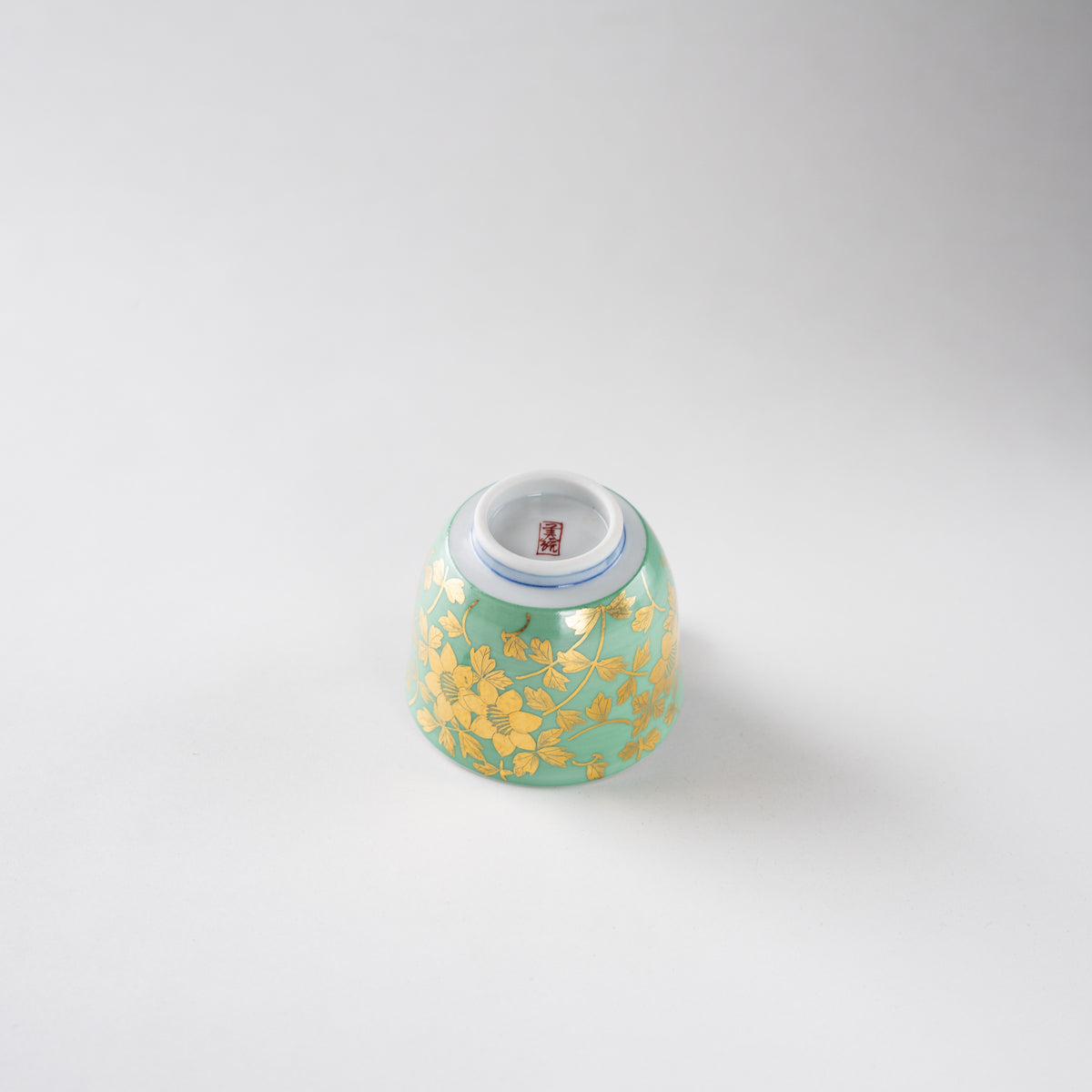 High-angle view of the inverted Kinrande floral arabesque GUINOMI sake cup by Mitsuru Yoshida, showing the circular foot ring, red square artist seal, and gilded patterns on a pale green ground.