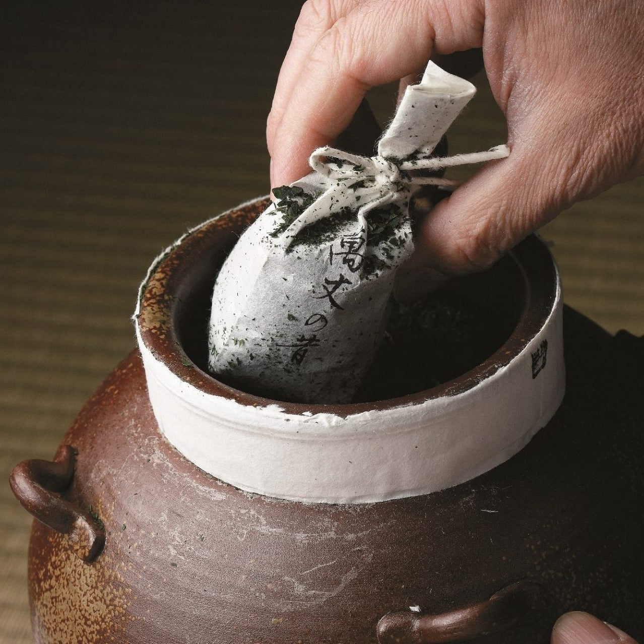 A close-up of an artisan's hand carefully retrieving a traditional paper-wrapped packet of tea leaves from a ceramic tea jar (chatsubo).