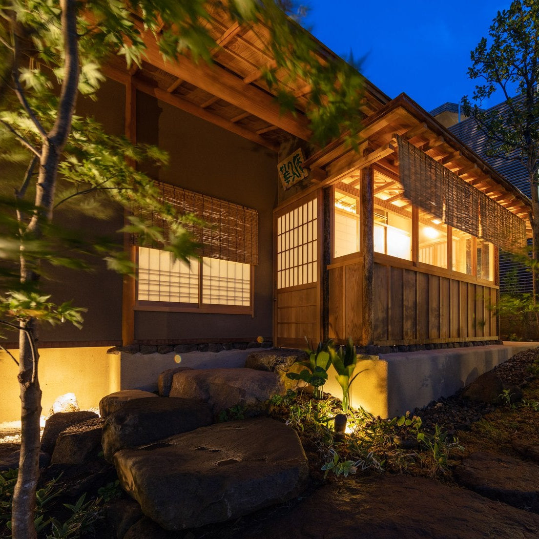 Wooden Japanese tea house illuminated at blue hour, surrounded by a lit garden with trees and a traditional stone lantern.