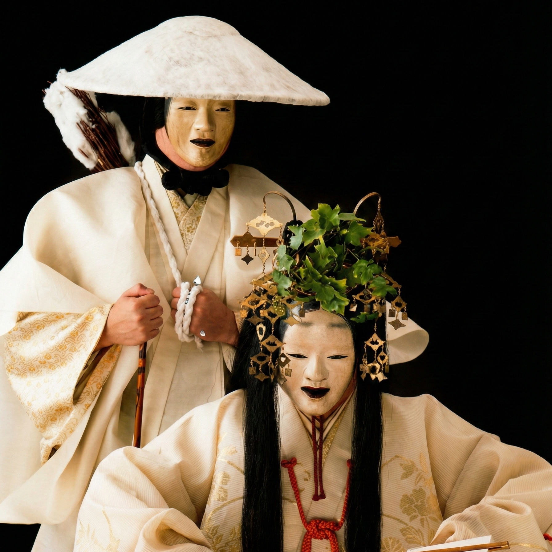 黒い背景の前に立つ、伝統的な白と金の衣装をまとった二人の日本の能楽師。前面の人物は緑の葉で飾られた精巧な金の頭飾りをつけ、黒い唇の面をかぶり、幣（ぬさ）を手に持っています。後ろに立つ人物は幅広い円錐形の白い笠をかぶり、面をつけて杖を持っています。