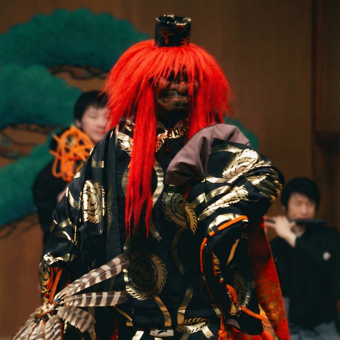 能舞台に立つ日本の能楽師。長く鮮やかな赤いかつらと面をつけています。衣装は、金の丸紋が入った黒の着物に、鮮やかな橙赤色の柄入り大口袴を合わせたものです。能楽師は羽根で作られた伝統的な扇を手に持ちます。背景では、松の木が描かれた伝統的な鏡板の前で、囃子方が笛を演奏しています。