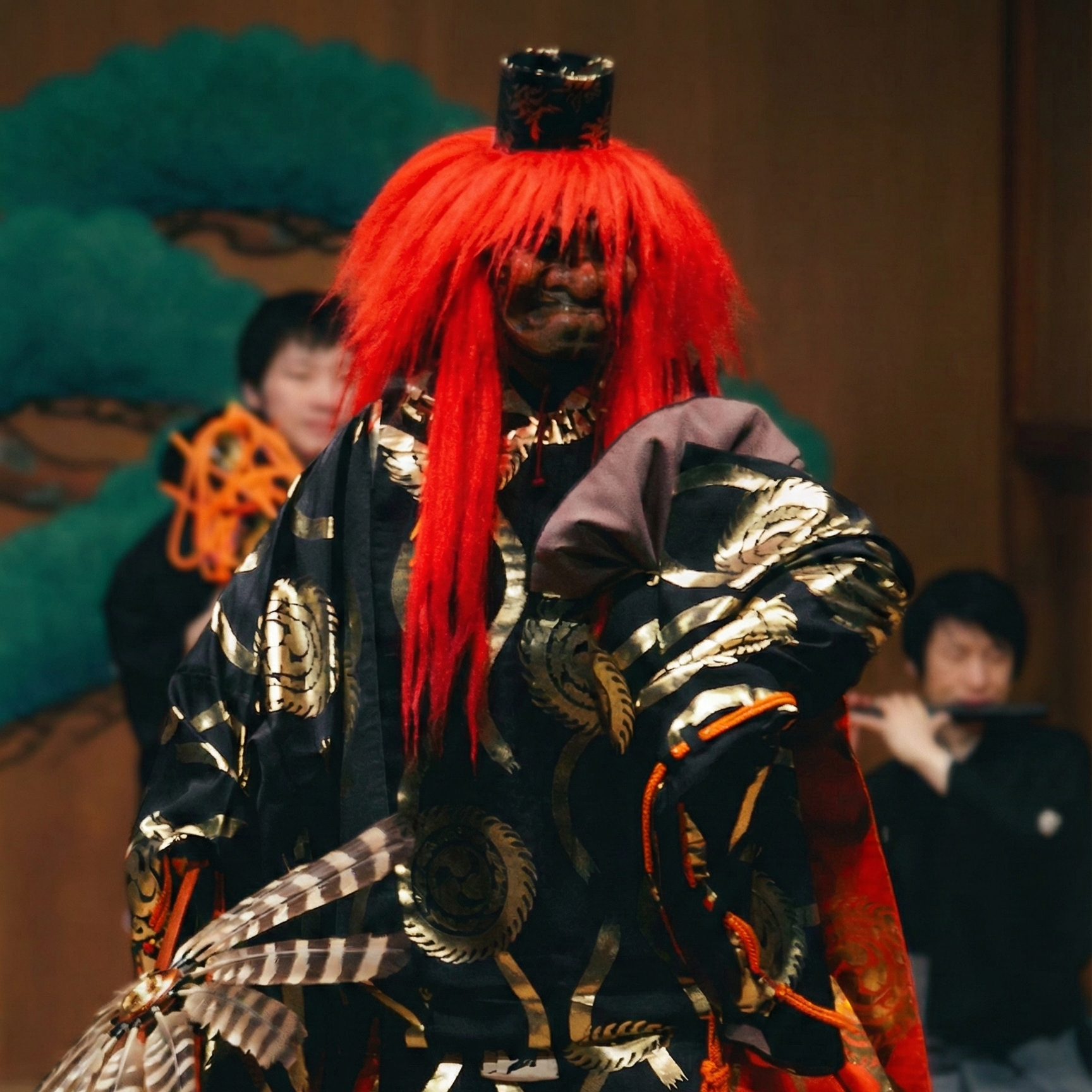 能舞台に立つ日本の能楽師。長く鮮やかな赤いかつらと面をつけています。衣装は、金の丸紋が入った黒の着物に、鮮やかな橙赤色の柄入り大口袴を合わせたものです。能楽師は羽根で作られた伝統的な扇を手に持ちます。背景では、松の木が描かれた伝統的な鏡板の前で、囃子方が笛を演奏しています。