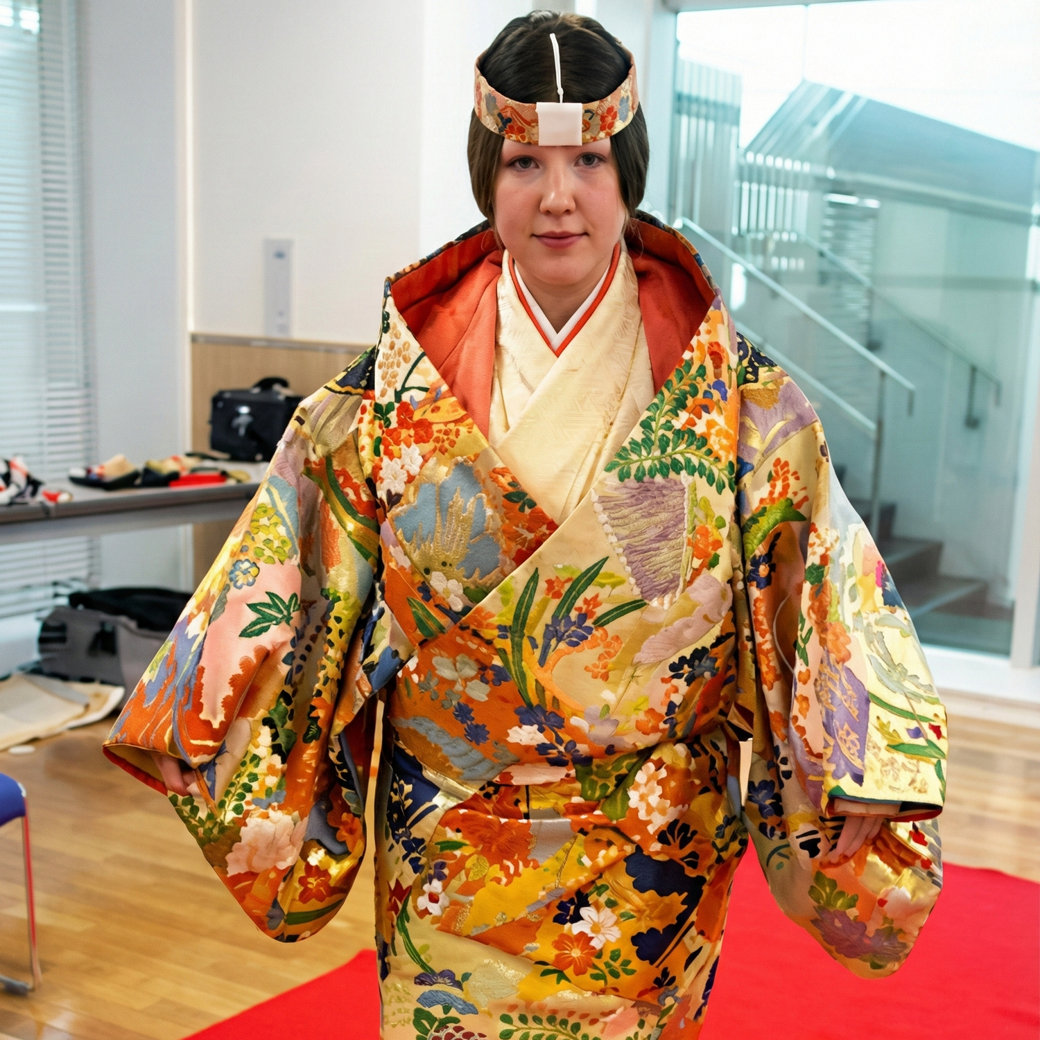 A Japanese Noh performer in a dynamic pose on stage, wearing a long, vibrant red wig and a mask. The costume consists of a black robe with large gold circular crests over bright orange-red trousers decorated with gold patterns. The performer holds a feathered fan. In the background, a musician plays a flute against a traditional pine tree backdrop.