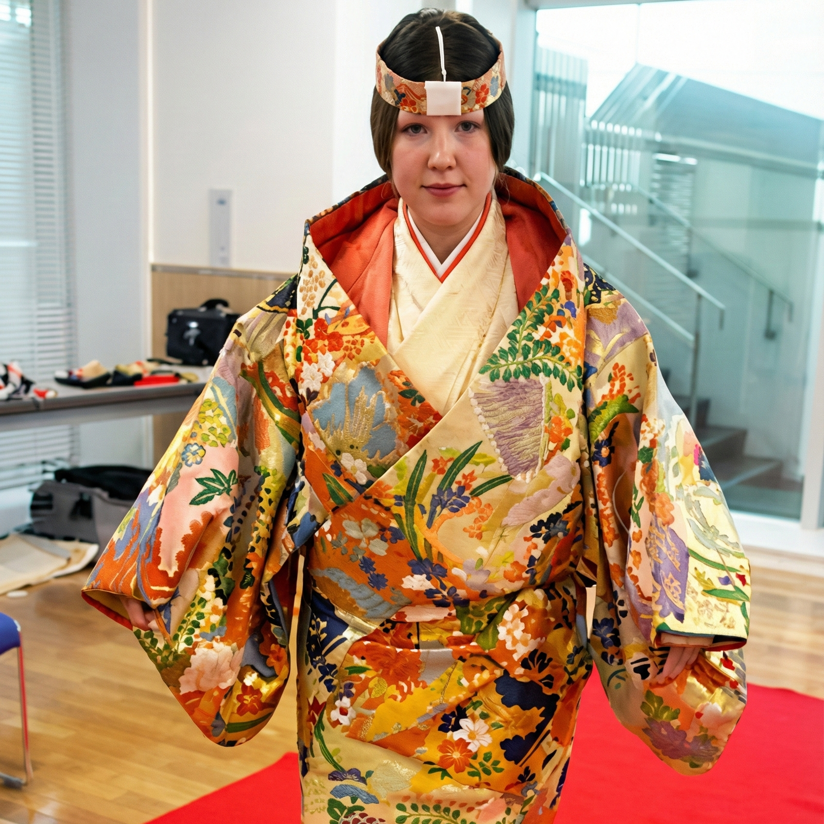能舞台で躍動的なポーズをとる日本の能楽師。長く鮮やかな赤いかつらと面をつけています。衣装は、金の大きな丸い紋様が入った黒の着物に、金模様で飾られた鮮やかな橙赤色の袴を合わせたものです。能楽師は羽根扇を手に持っています。背景では、松の木が描かれた伝統的な鏡板の前で、囃子方が笛を演奏しています。
