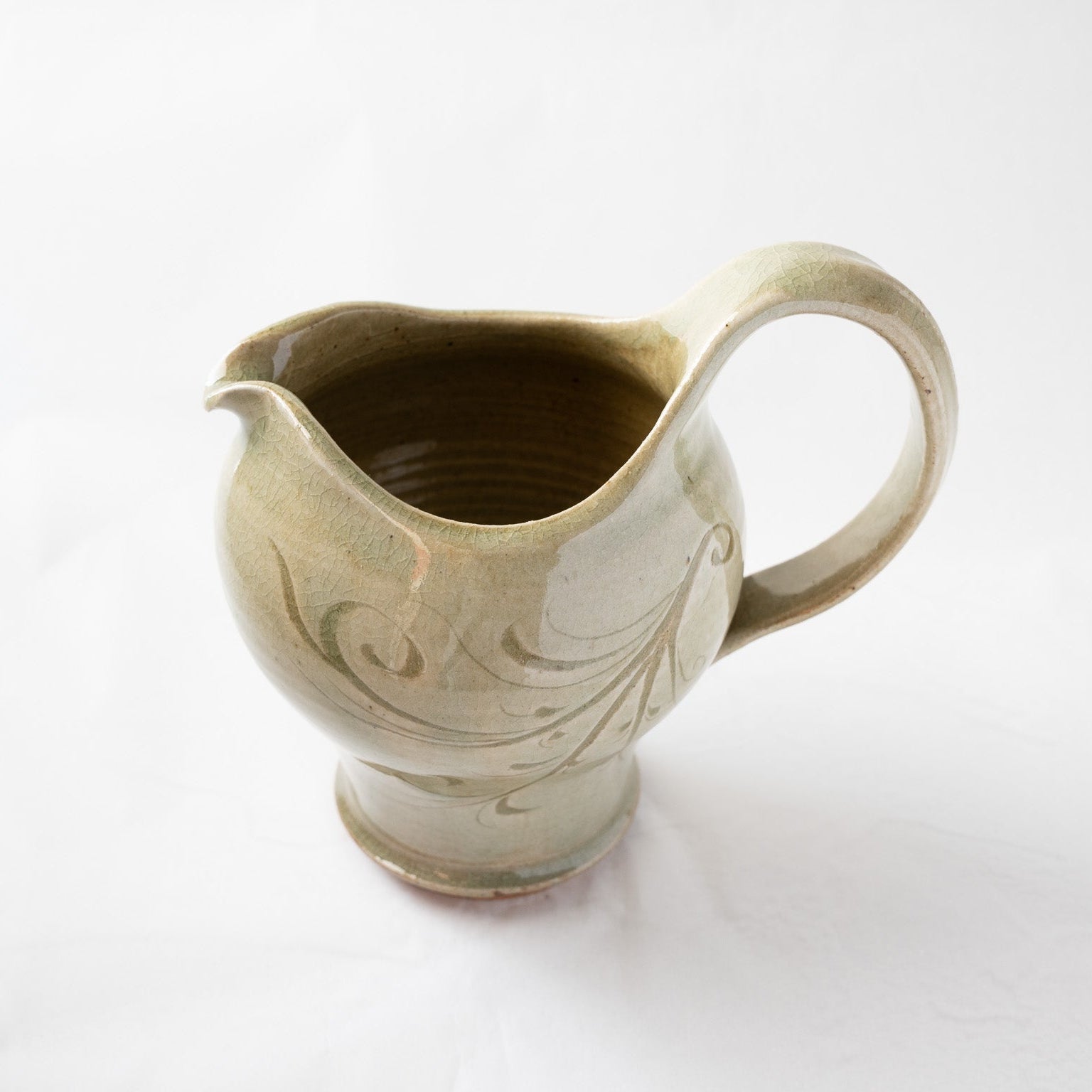 An elevated, three-quarter view of an ash-green glazed ceramic pitcher by Toyo Kobayashi, highlighting the smooth inner rim, delicate spout, and fluid handle line.