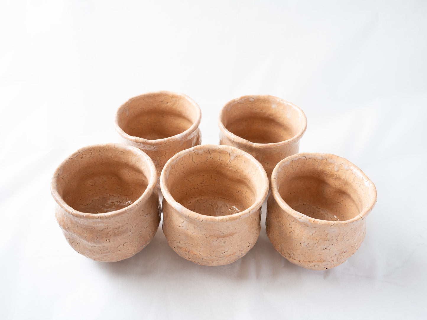 Front view of the five-piece "Satoshi" cup set arranged together, illustrating the organic variations in form, lip shapes, and soft light reflections on the glazed surfaces.