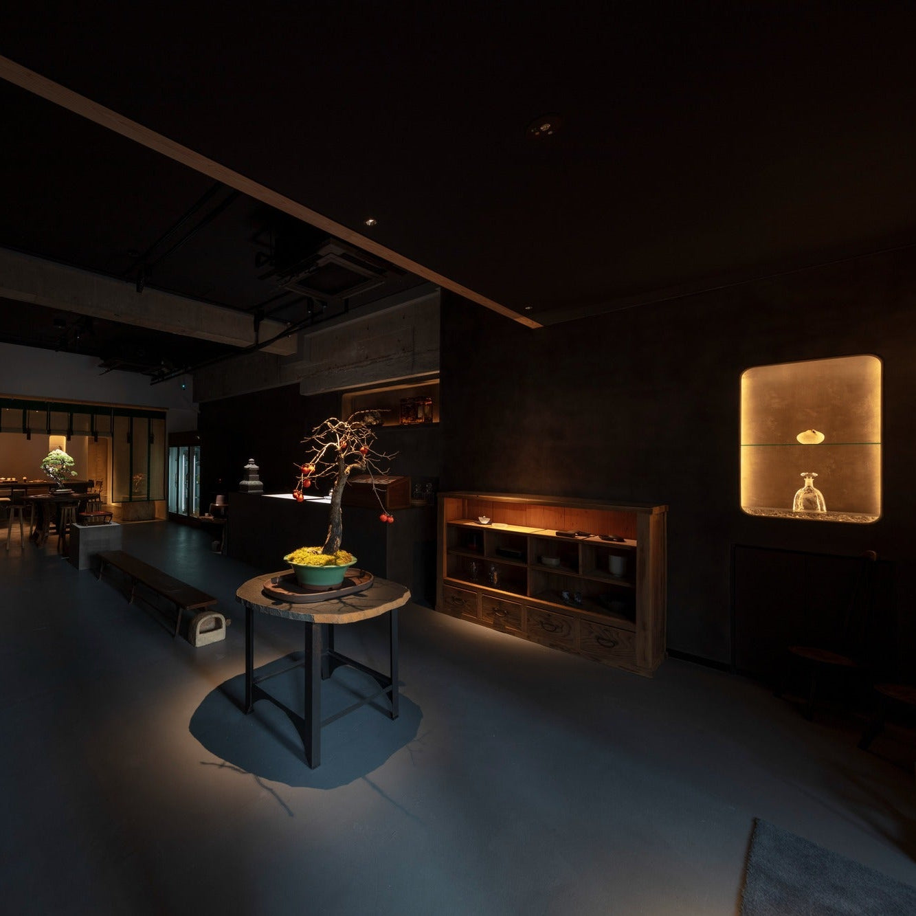A dim room of bonsai on stands and tables opens toward a glowing counter beyond, restrained Japanese architecture.