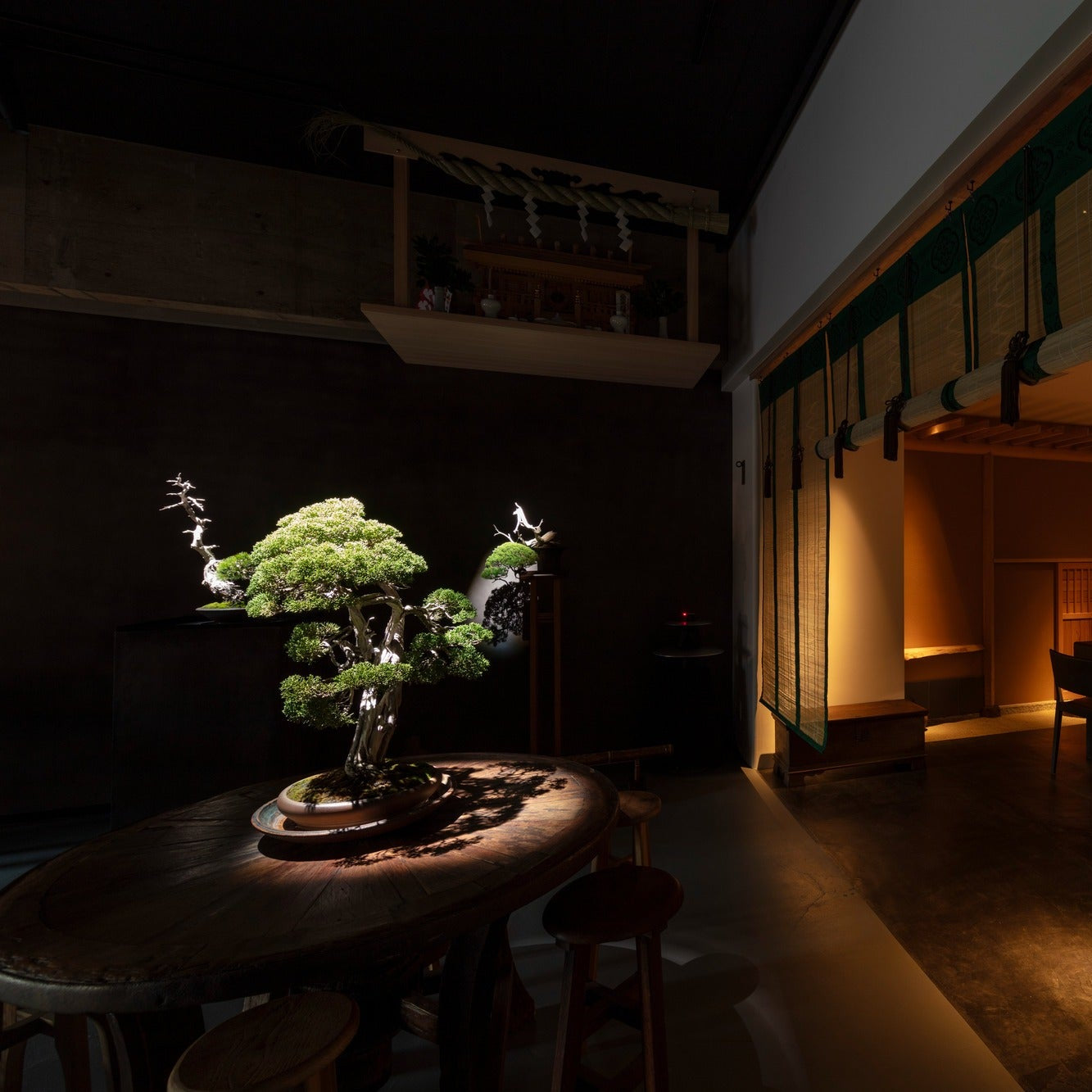 A bonsai on a round table below a small Shinto shelf hung with a sacred rope and paper streamers.