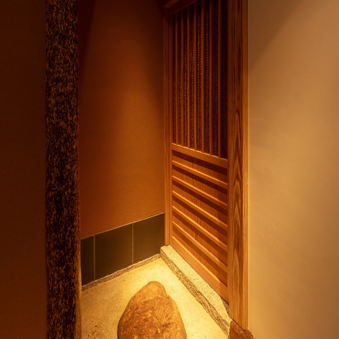 A rough stone stands on raked sand in a corner where a granite post meets a cedar lattice screen.