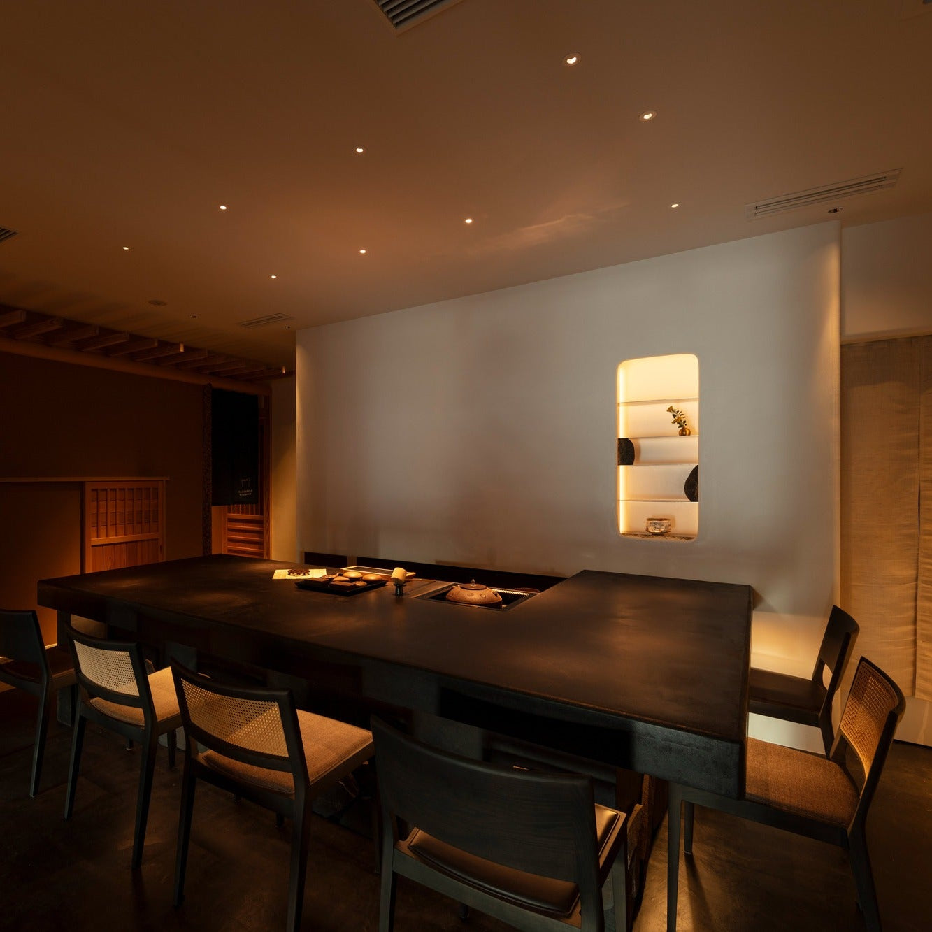 A long dark counter with caned chairs and a lit niche of tea bowls anchors the japanese cafe.