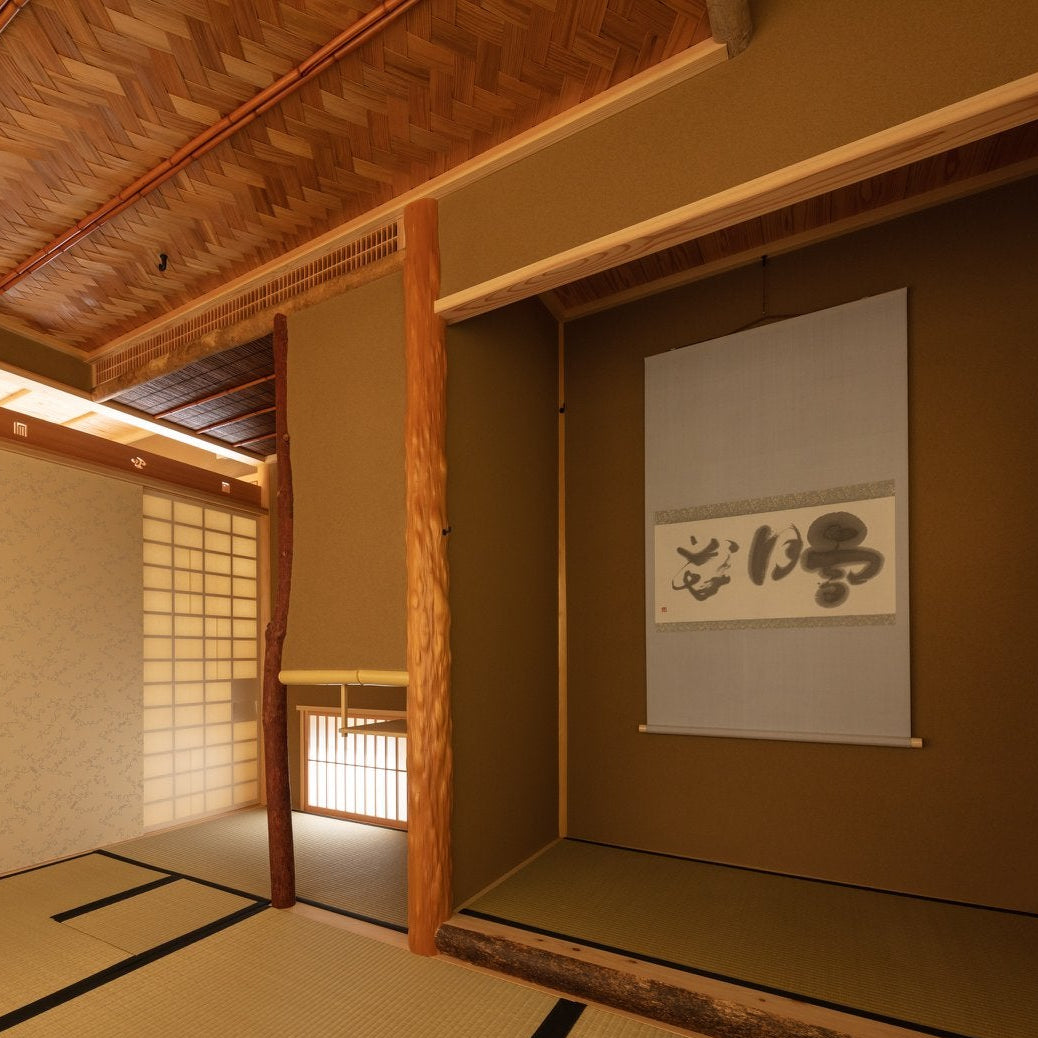 Japanese tea room with a tokonoma alcove and calligraphy scroll beneath a woven bamboo ceiling, traditional chashitsu architecture.