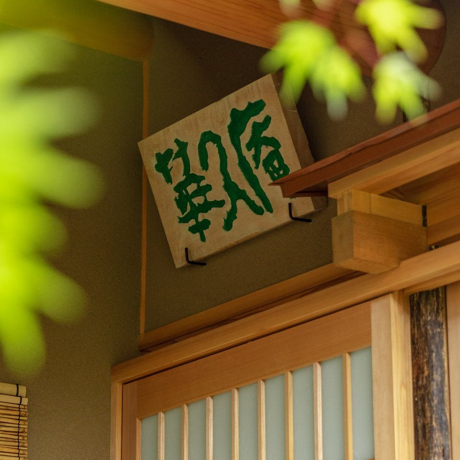 Hand-carved wooden name plaque framed by green maple leaves, a detail of the Japanese tea room.