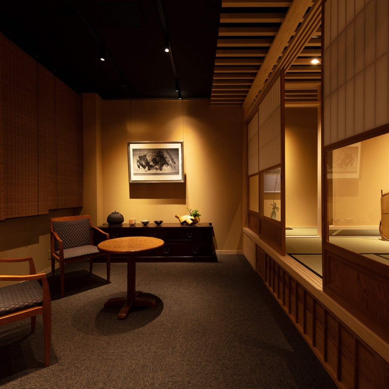 Waiting area framed by reed blinds with armchairs and a round table beside a shoji-lit tea room.