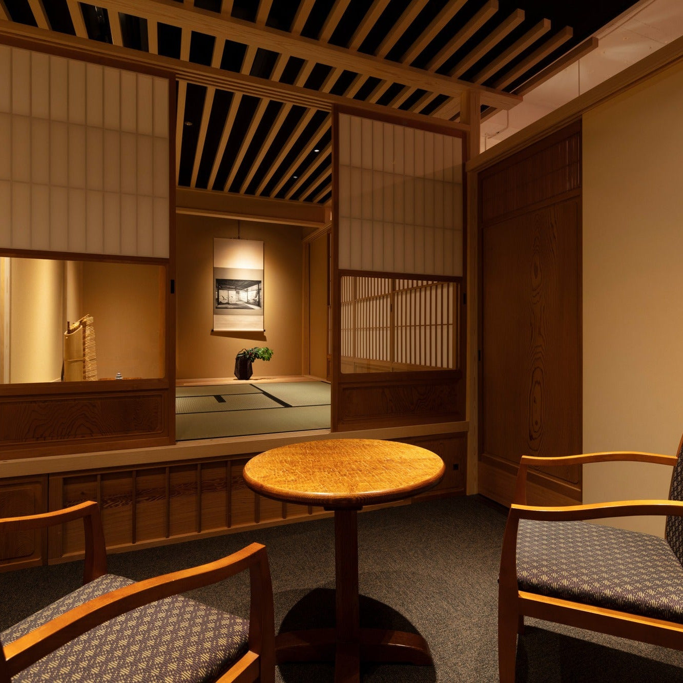 Waiting area with a round table and armchairs looking into a tatami tea room with a hanging scroll.