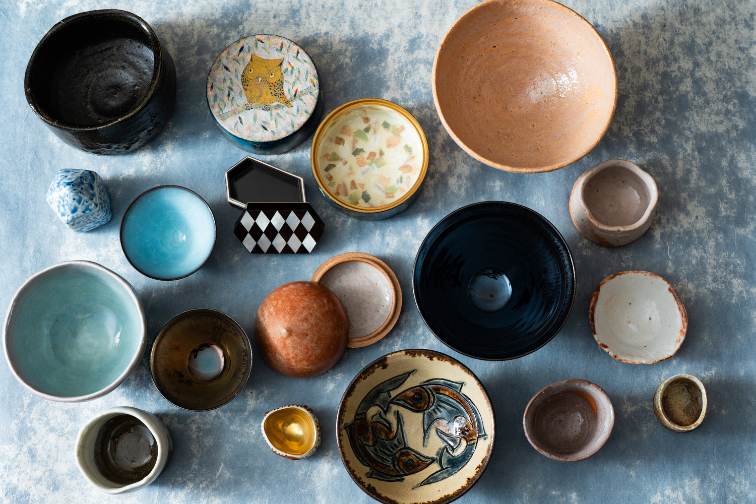 A curated collection of diverse contemporary Japanese ceramic vessels and artisanal objects, including chawan and kogo, arranged top-down on a textured cerulean background. The arrangement showcases the profound variation in glaze, form, and texture—from raw earthiness to refined porcelain—symbolizing the breadth and freedom of modern Japanese craft and the spirit of 'wabi' in a contemporary context.