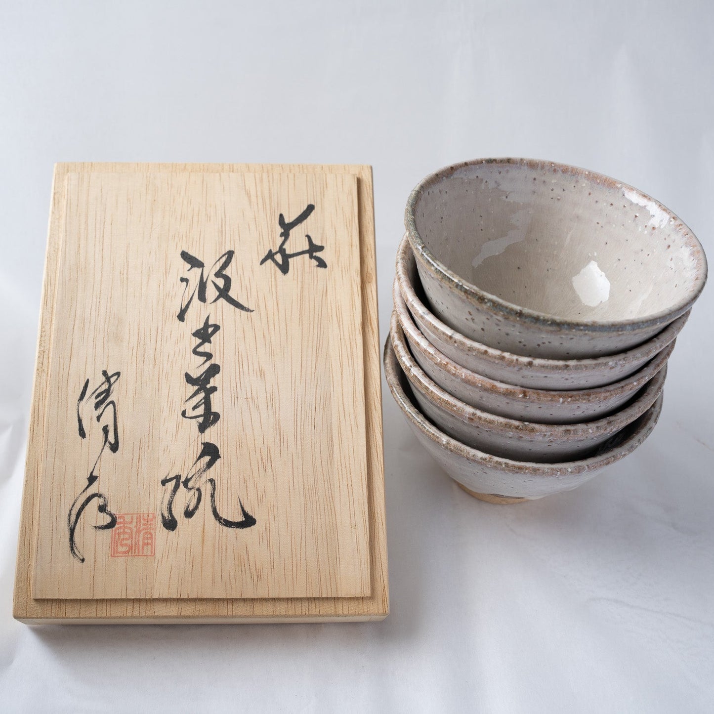 A vertical stack of five Hagiyaki tea cups placed next to their traditional paulownia wooden box with handwritten Japanese calligraphy and a red artist stamp.