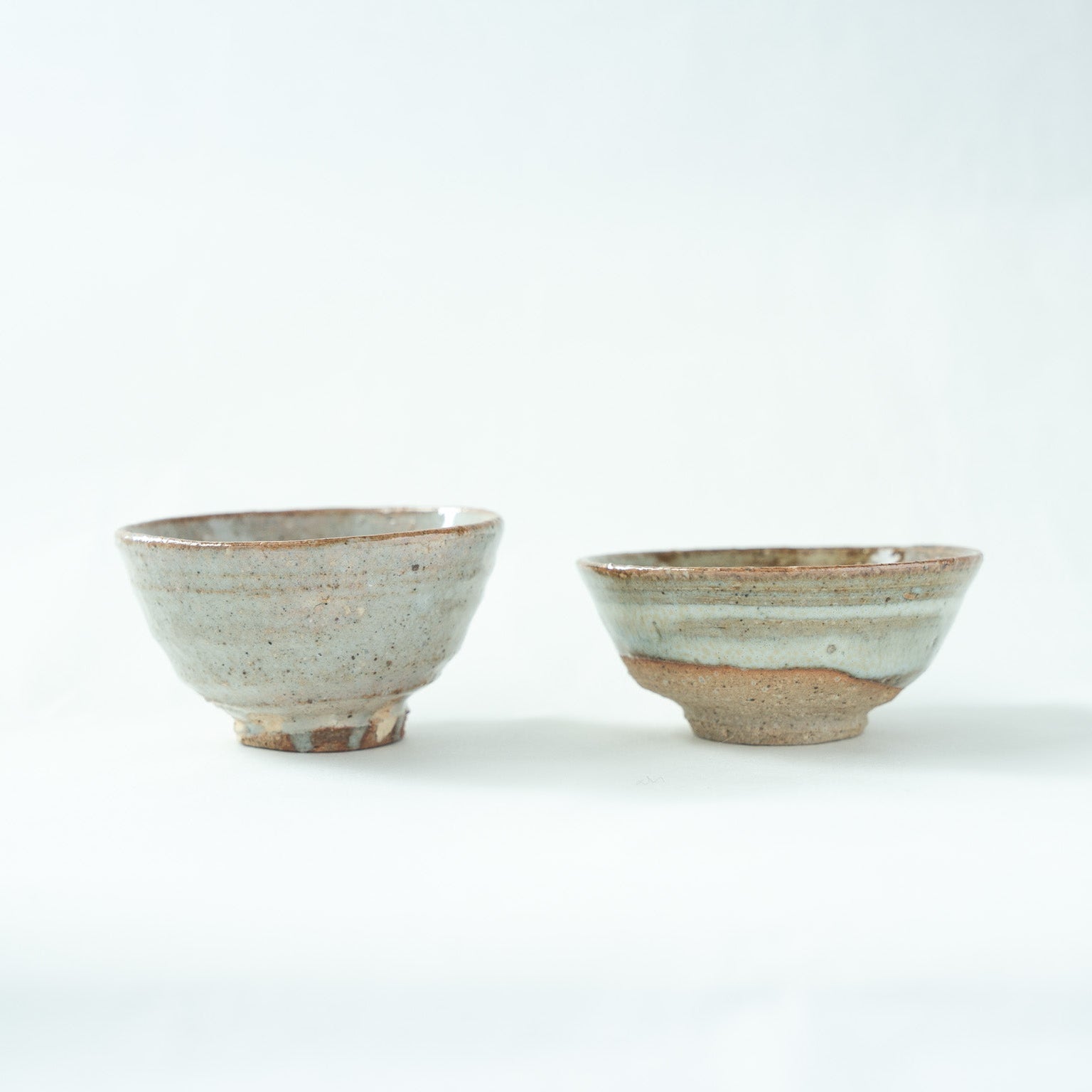 Front view of two soft blue-gray GUINOMI by Hajime Kato side by side, showing a slightly deeper cup on the left and a wider, shallow bowl on the right.