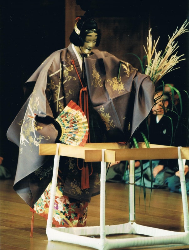 A Japanese Noh performer portraying a female character, wearing a white mask and an elaborate costume featuring a muted purple outer robe with gold floral patterns over a vibrant, colorful floral kimono. The performer holds a decorative folding fan and stands next to a wooden stage prop representing a simple structure, adorned with tall pampas grass.