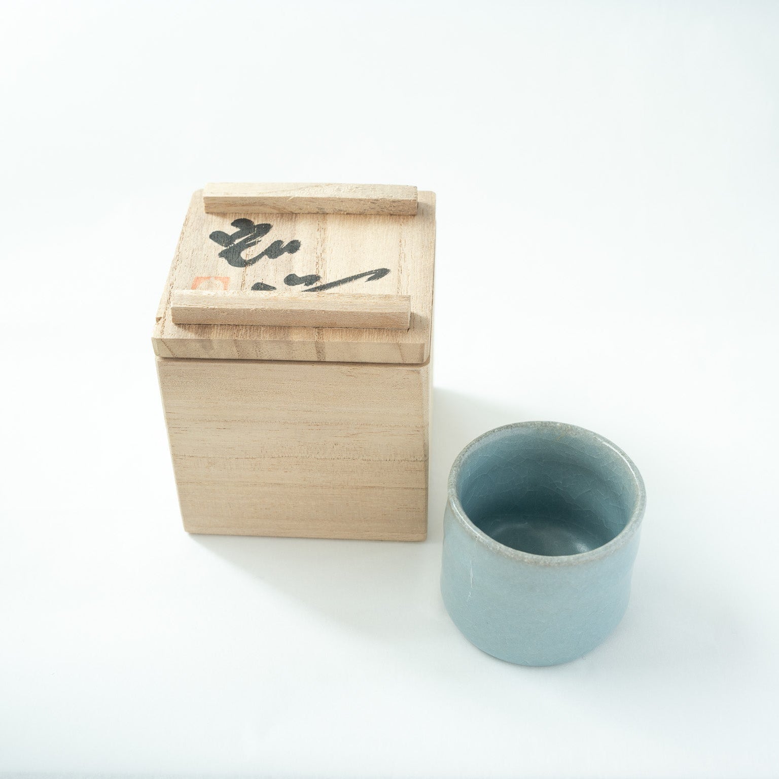 The celadon sake cup shown with the wooden box lid inverted, revealing the artist's handwritten signature "Hiro" and red seal on the reverse side.