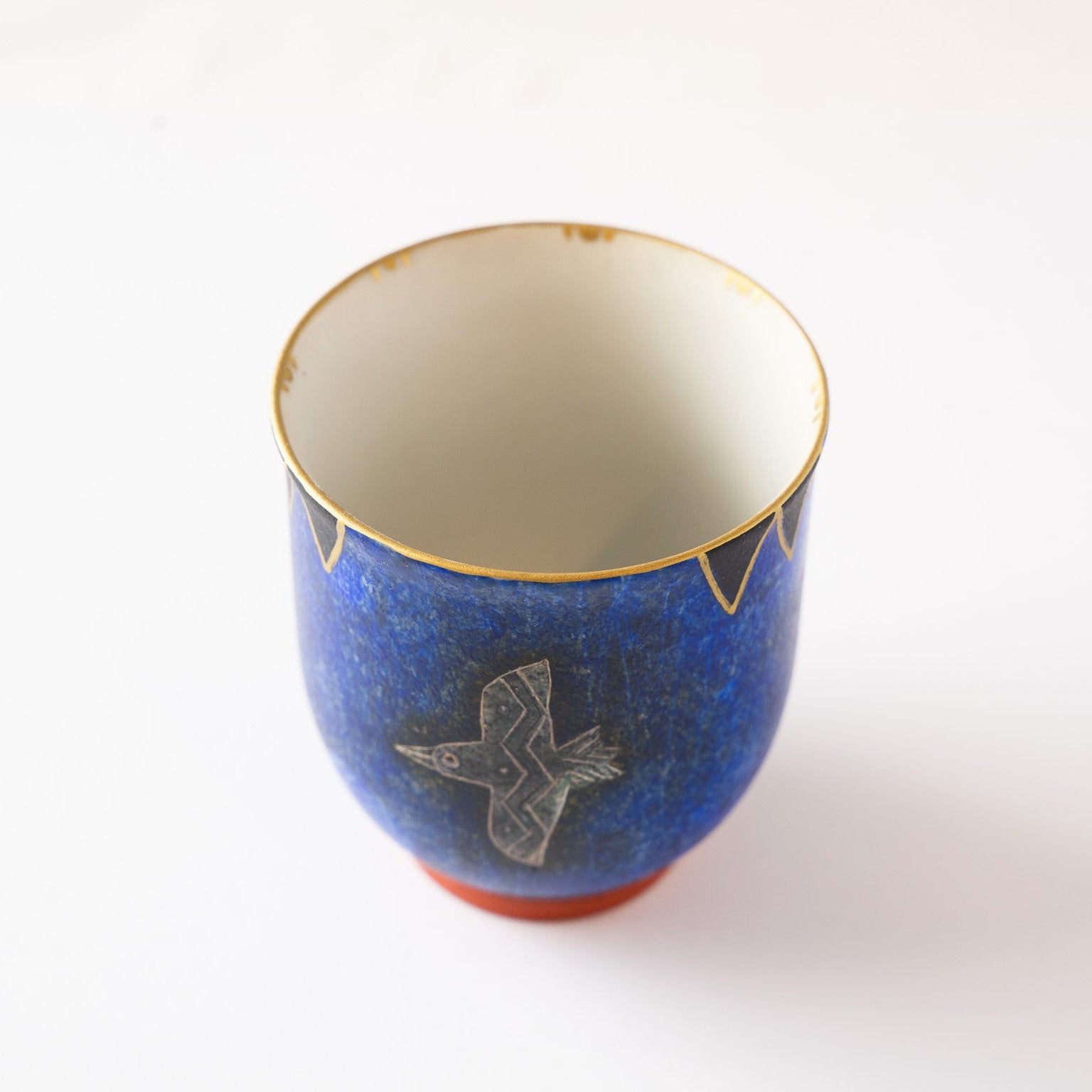 High-angle view of a deep blue ceramic YUNOMI cup featuring an engraved silver bird motif, white interior, and a gold-rimmed geometric pattern on a white background.