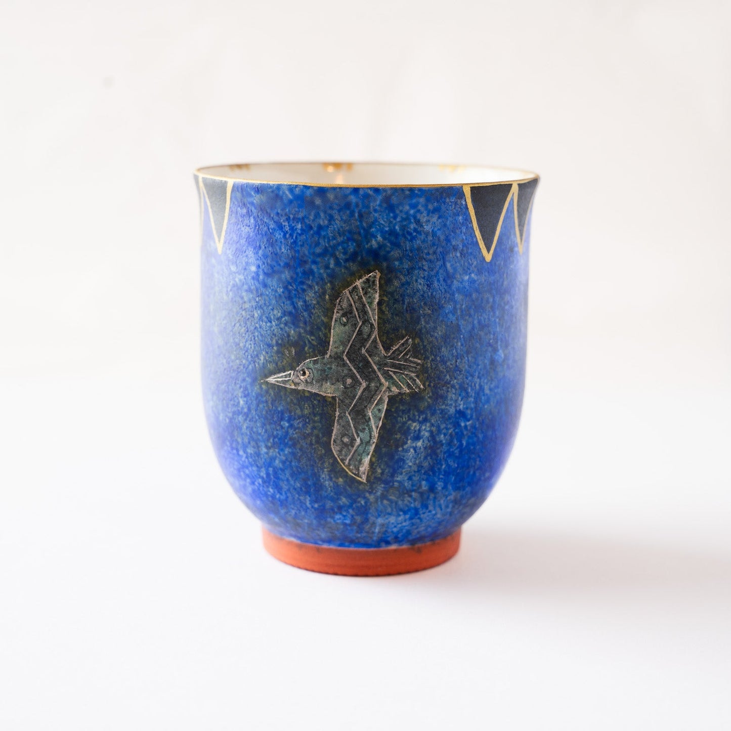 Slightly turned view of the ceramic cup demonstrating how natural light interacts with the silver enamel bird and the multi-layered indigo glaze.