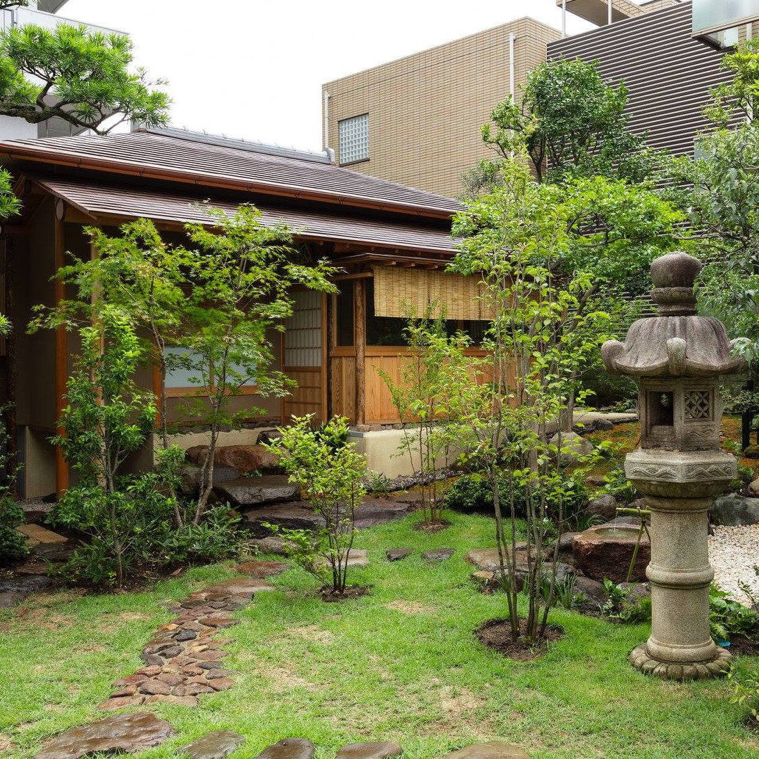 Entrance of a wooden tea house with a hanging wooden name plaque, viewed through bright green Japanese maple leaves in the foreground.