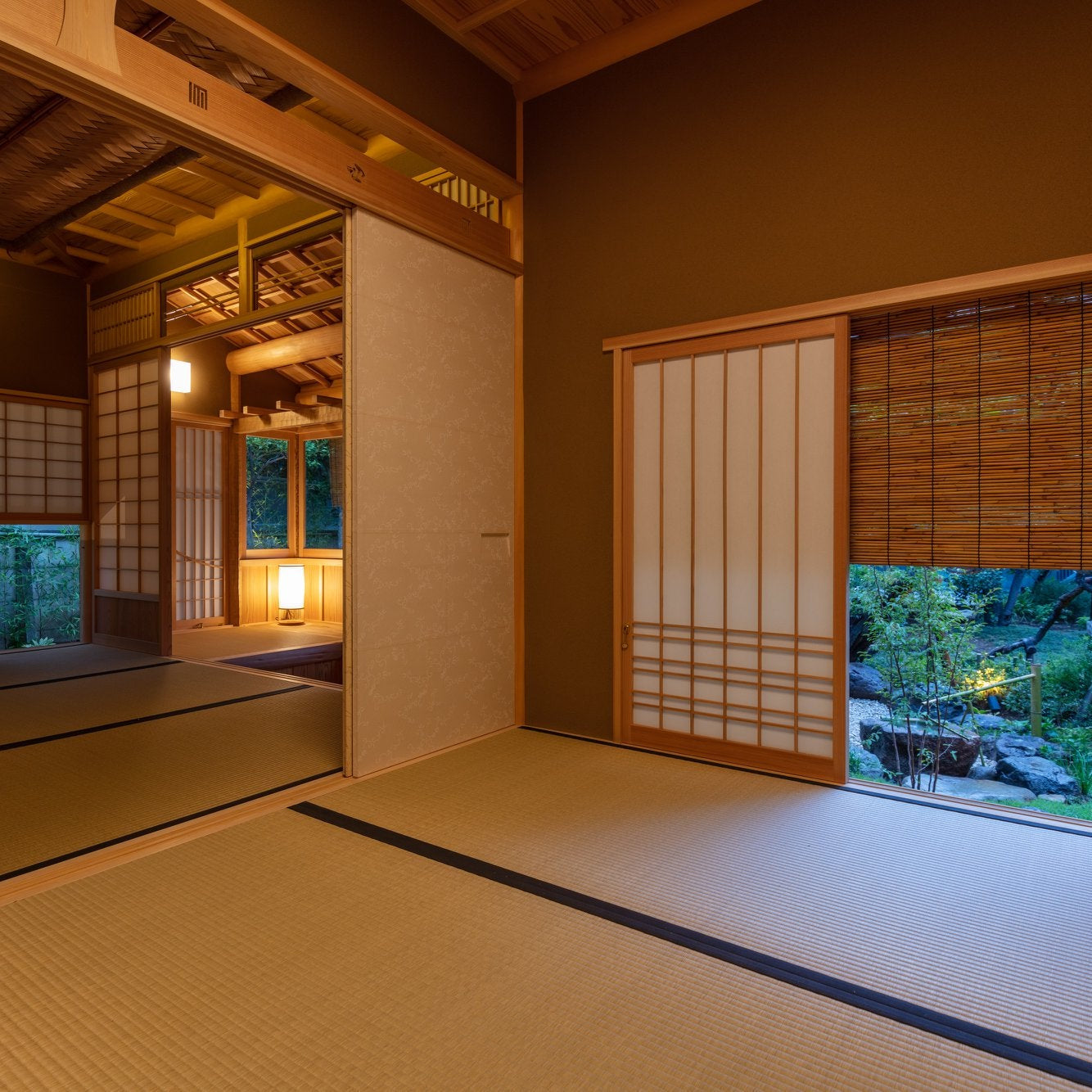Interior of a Japanese tea room with timber walls and a low bench, corner windows looking onto a green garden, and a lit washi andon lamp.