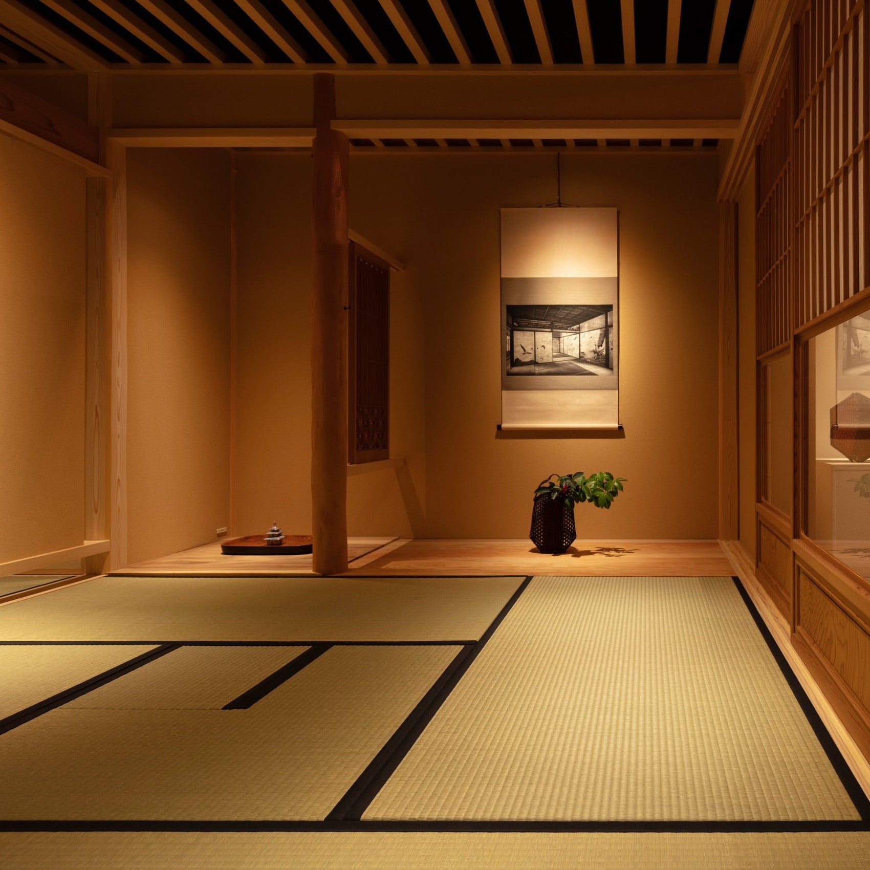 A serene traditional Japanese tearoom with warm lighting, featuring a tokonoma alcove with a hanging scroll, seasonal ikebana flower arrangement, and tatami mat flooring.