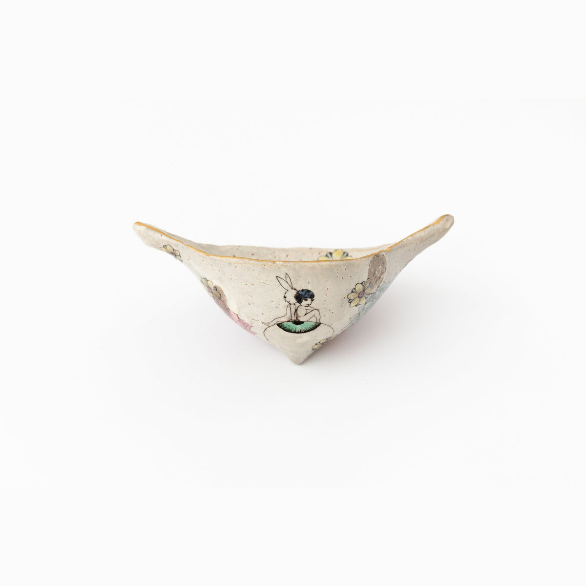 Front view of the small winged ceramic object on a white ground, a tiny figure and flowers painted on its pale surface.