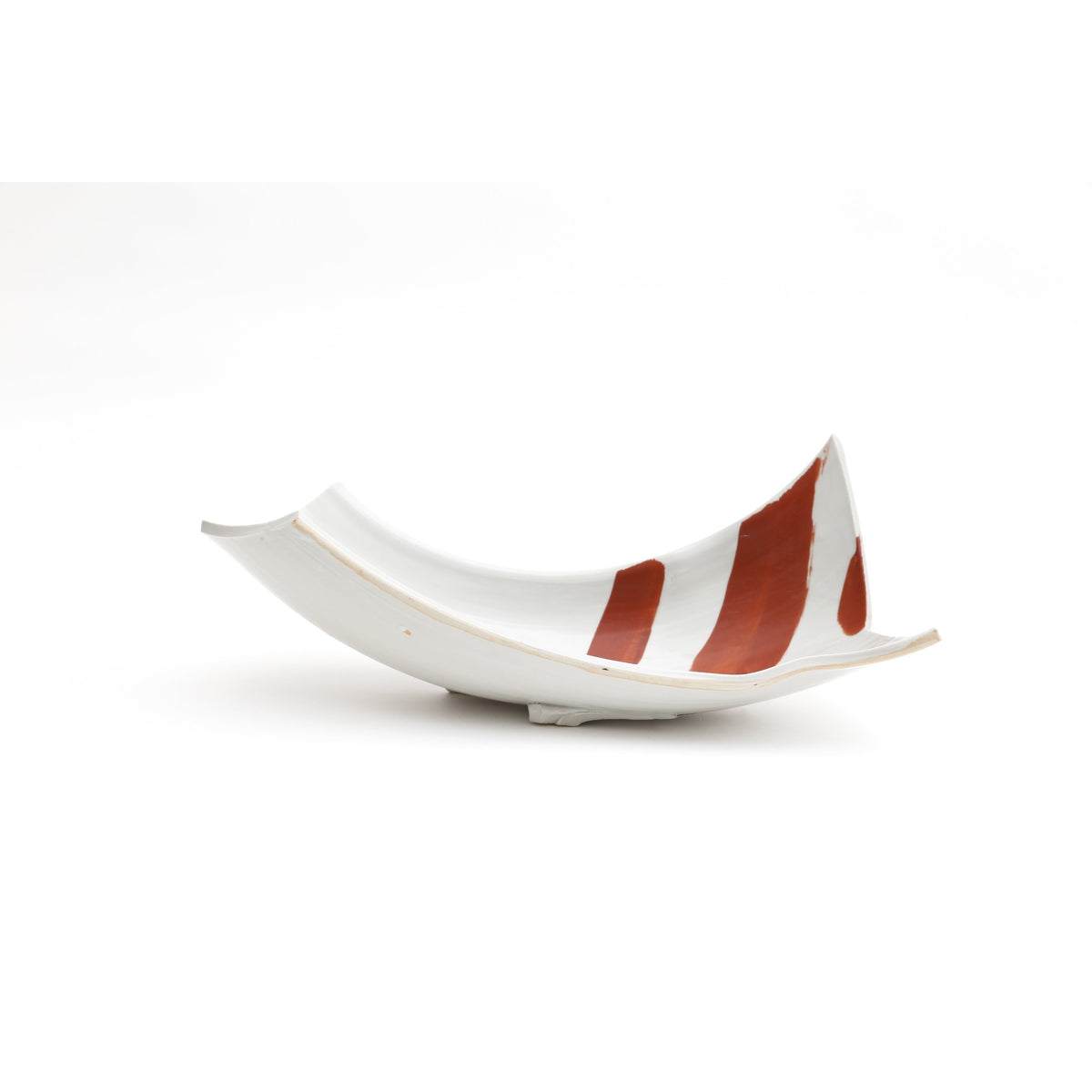 Vue de profil du long plat blanc sur pied, sur fond blanc, ses extrémités relevées et de larges bandes d'émail rouge traversant la surface.