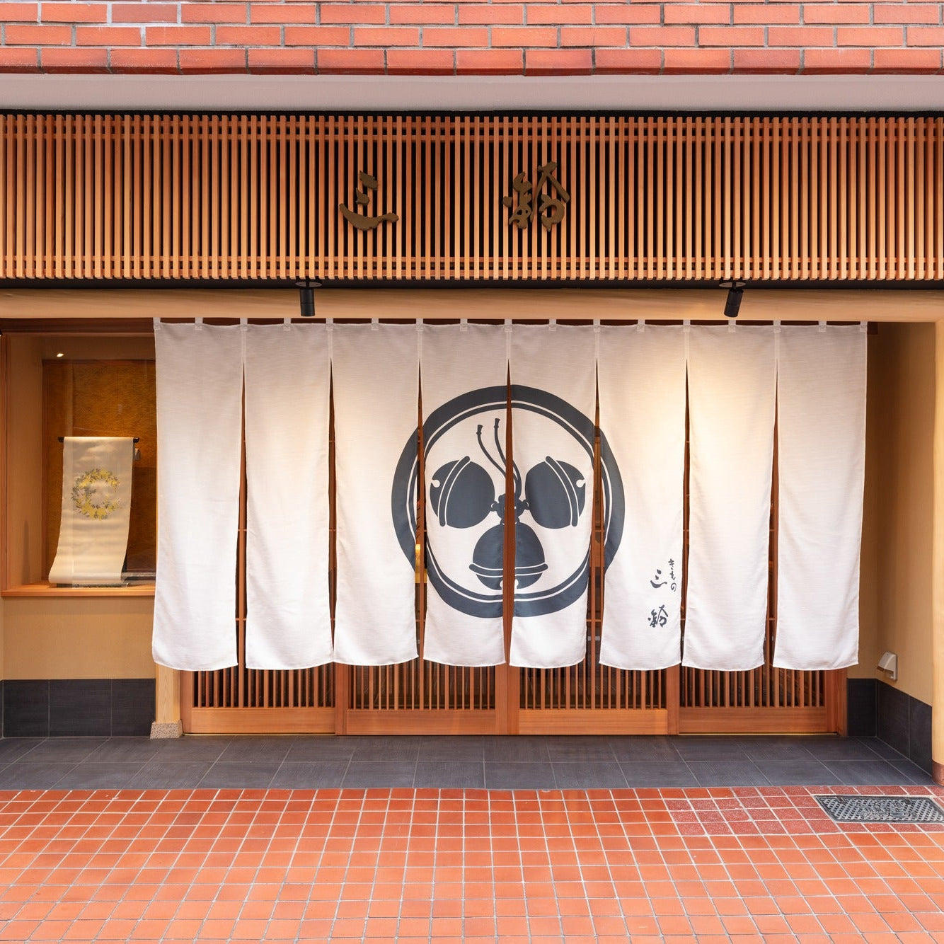 Brick-fronted shop with a slatted timber sign board and a white crested noren