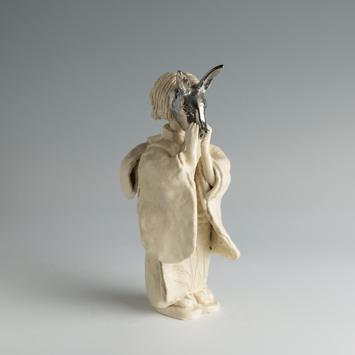Vue de trois quarts de la figure de jeune fille en grès clair au masque de renard sur la tête, photographiée sur fond gris de studio.