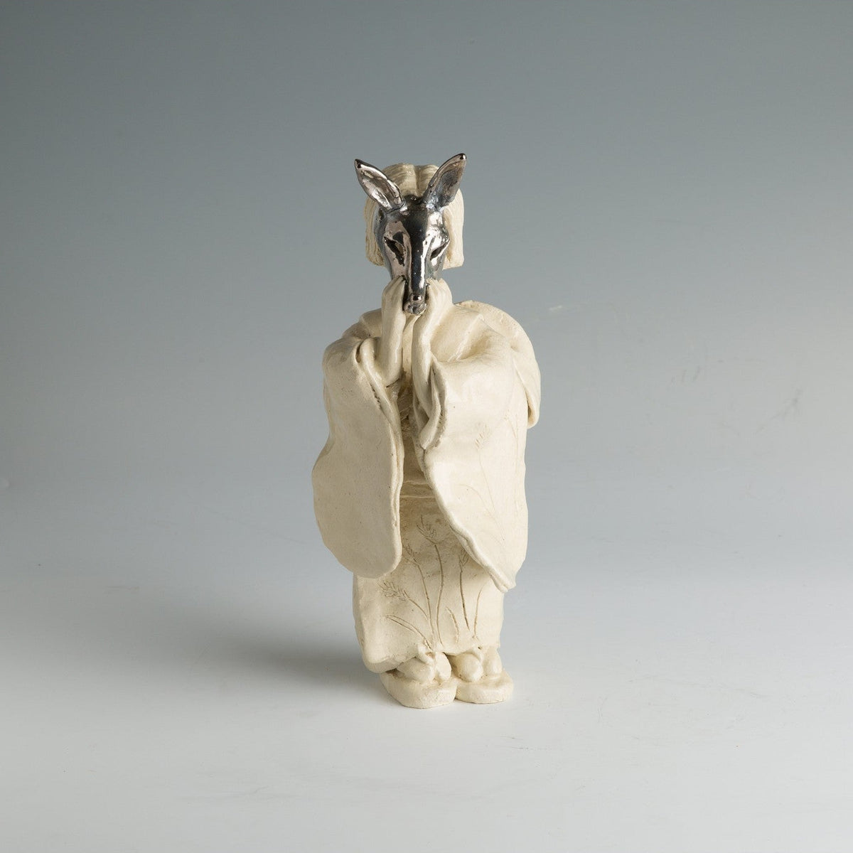 Vue de face de la figure, les manches du kimono rassemblées sur la poitrine et les oreilles du masque de renard dressées au-dessus de la tête.