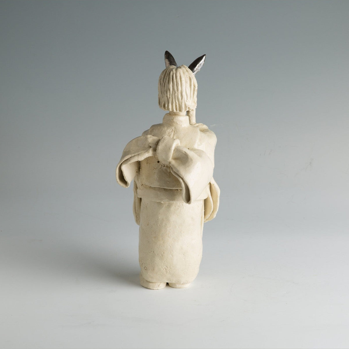 La figure vue sous l'autre angle de face, le visage baissé sous le masque relevé, en lumière grise douce.