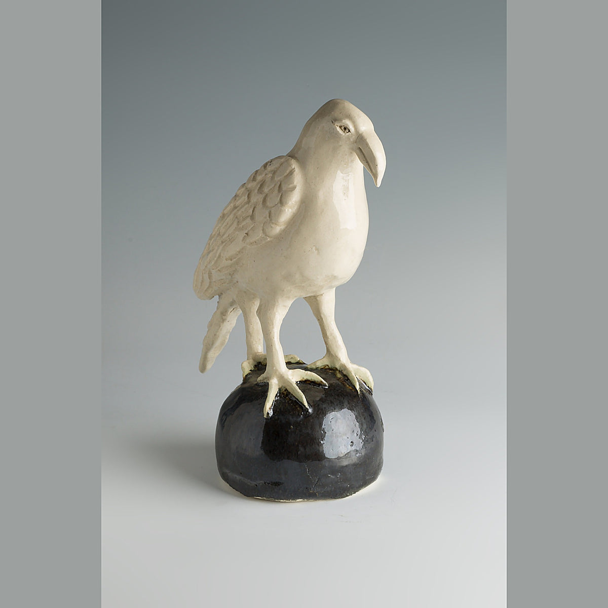 Front view of the pale stoneware crow sculpture standing on a rough black sphere, against a grey studio background.