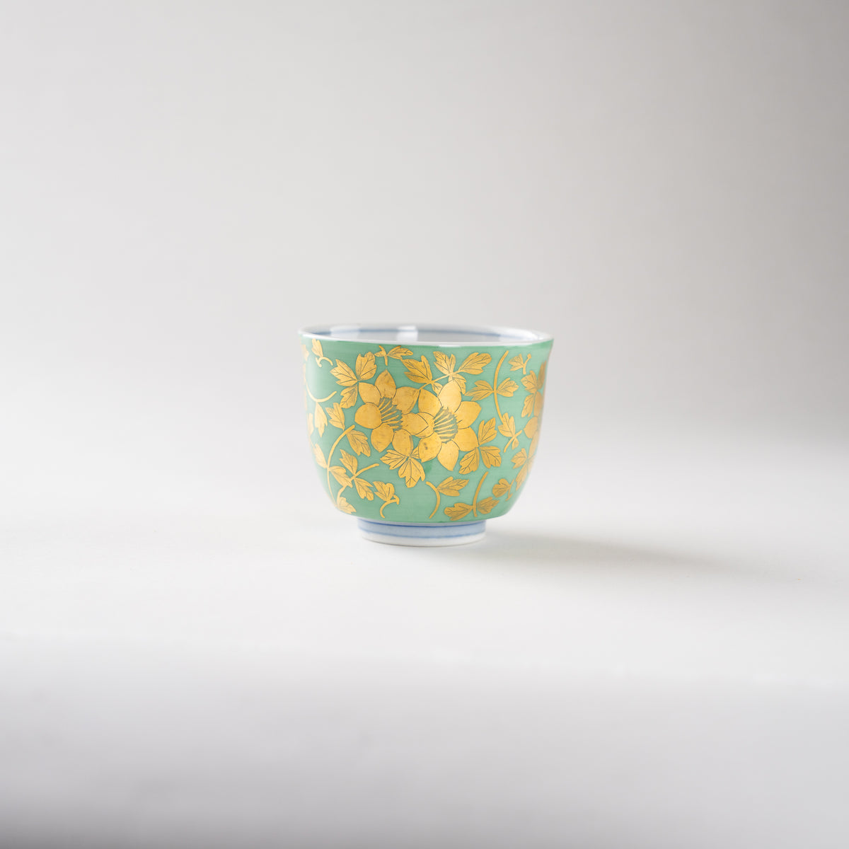 High-angle studio view of Mitsuru Yoshida's Kinrande GUINOMI sake cup, showcasing the intricate gold floral arabesque painted on a pale green ground.
