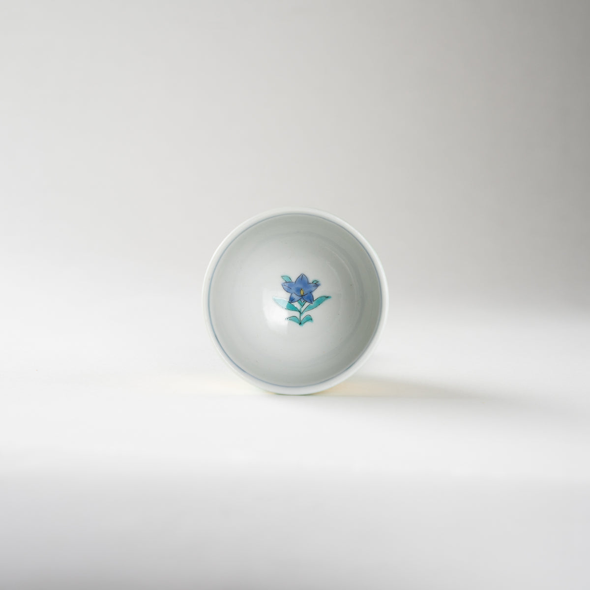 Top-down interior view of Mitsuru Yoshida's GUINOMI sake cup, revealing a single painted cobalt blue and green blossom at the center of the white basin.