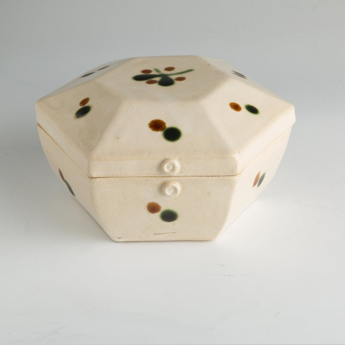 Front view of the ivory hexagonal lidded box on a grey ground, green and brown glaze dots on its panels.
