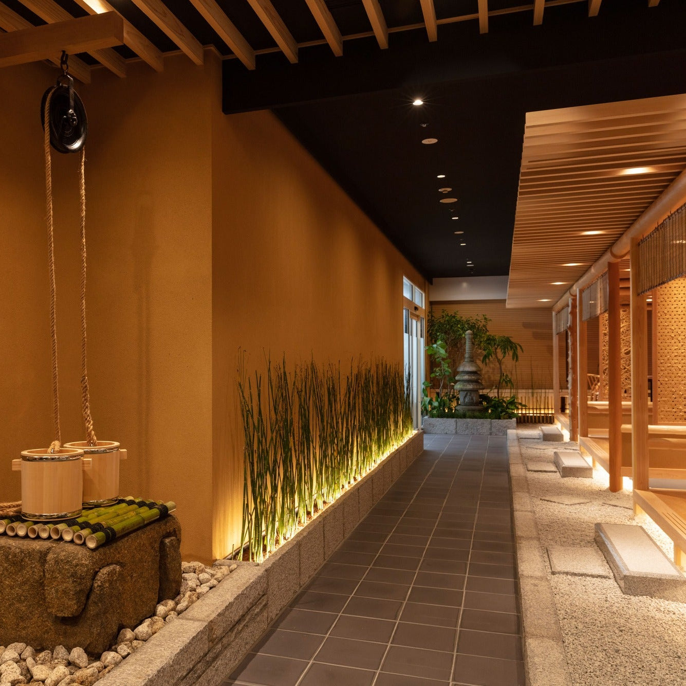 A stone well with rope pulley and wooden buckets sits along a corridor lined with horsetail, a stone pagoda beyond.