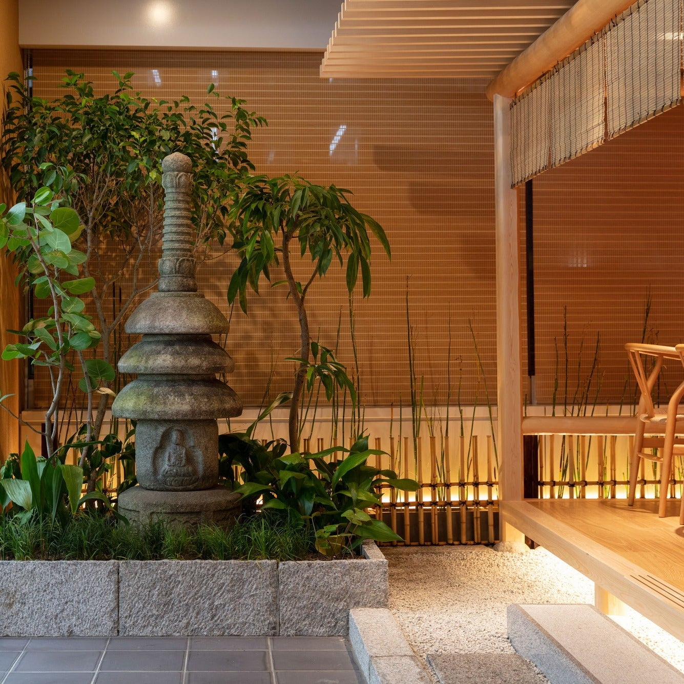 A tiered stone pagoda stands among greenery in a planted courtyard, beside a glazed wall and a reed blind.