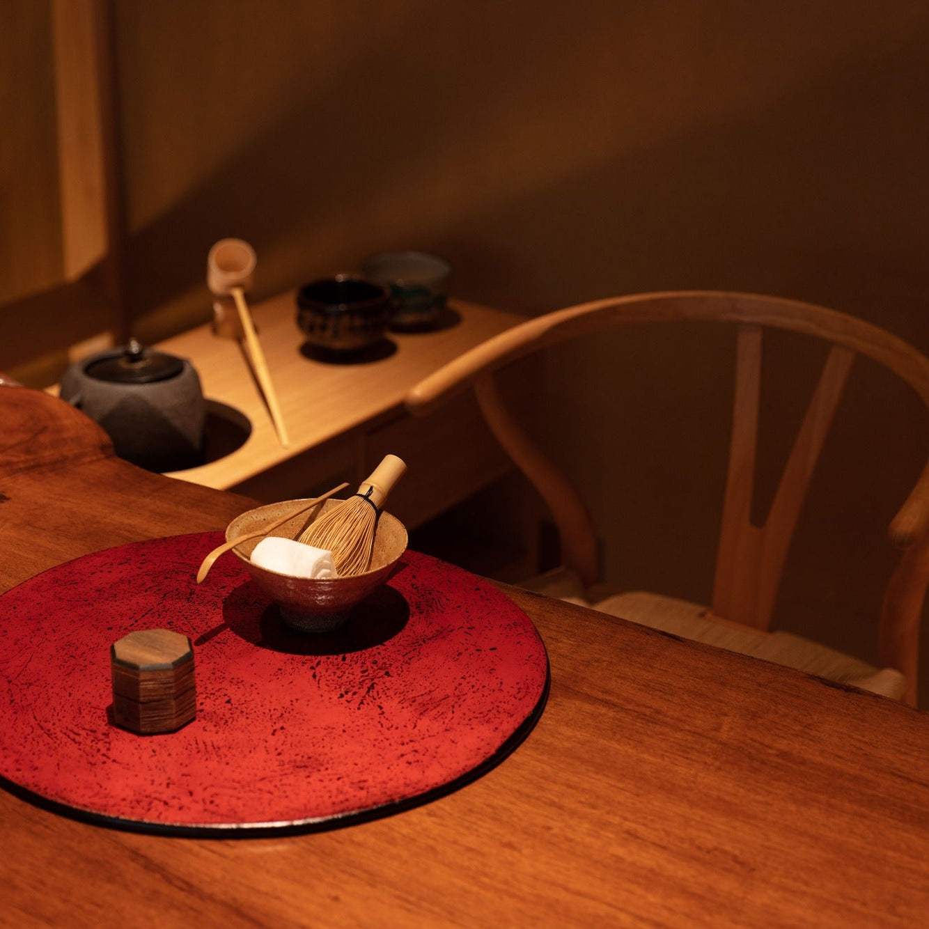 A tea bowl and bamboo whisk rest on a red lacquer tray, with a kettle and ladle set for matcha at the chashitsu.
