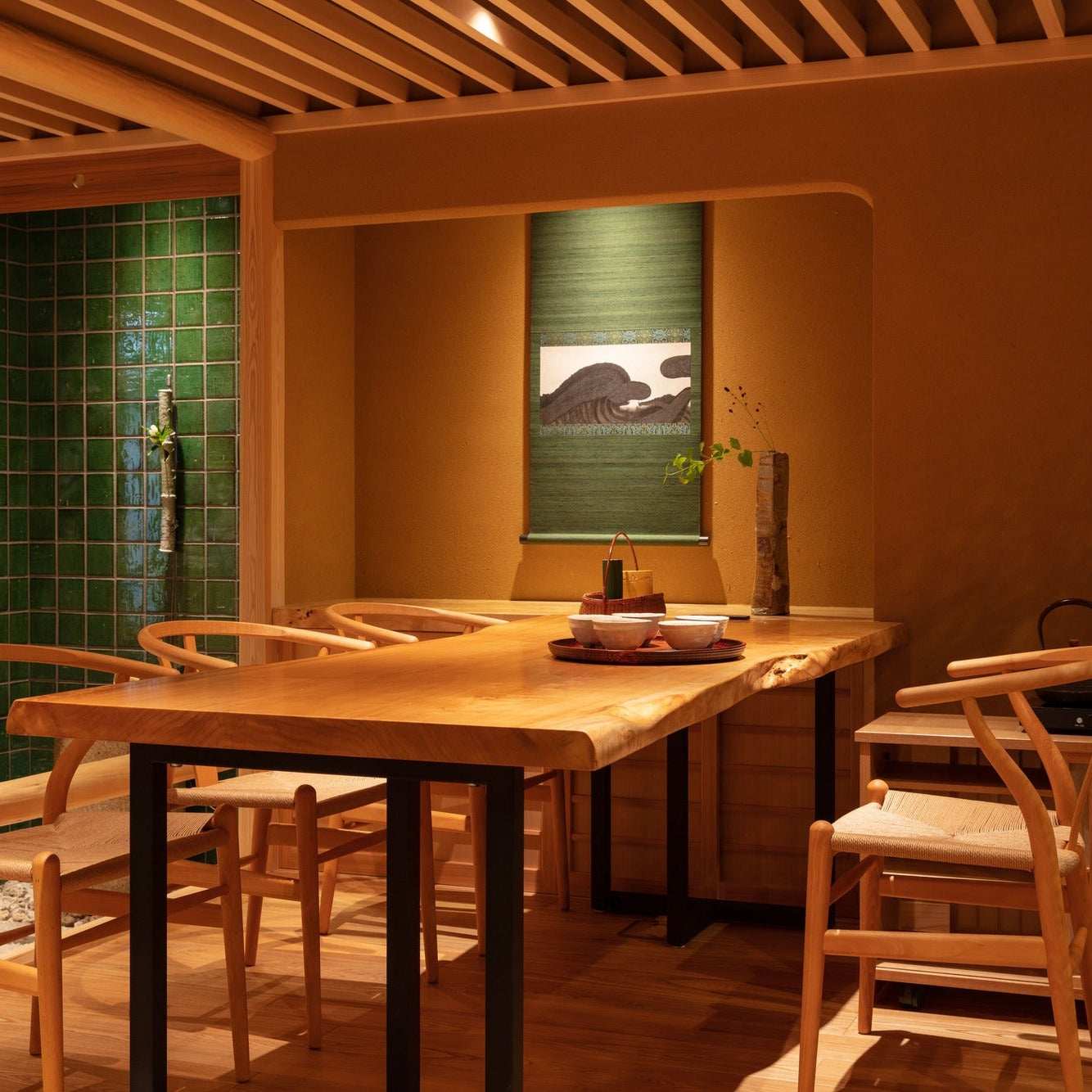 A live-edge table and woven-paper chairs before a green-tiled wall and a hanging scroll, a quiet corner of the Japanese office.
