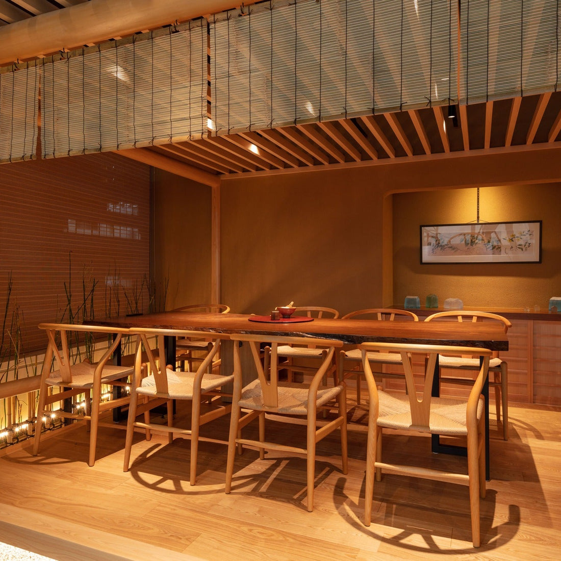 Woven-paper chairs at a live-edge table beneath a rolled reed blind, with horsetail planting lit along the platform edge.
