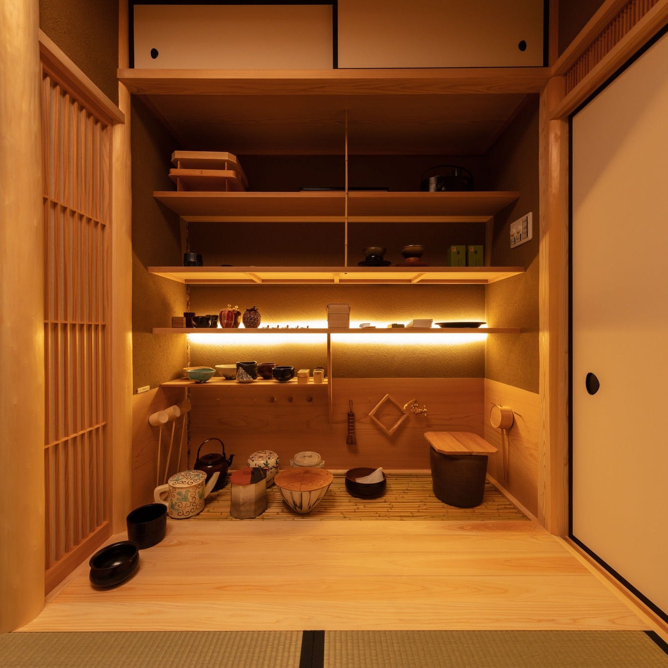 A mizuya preparation alcove lined with shelves of tea bowls, ceramics and bamboo tools under soft light.