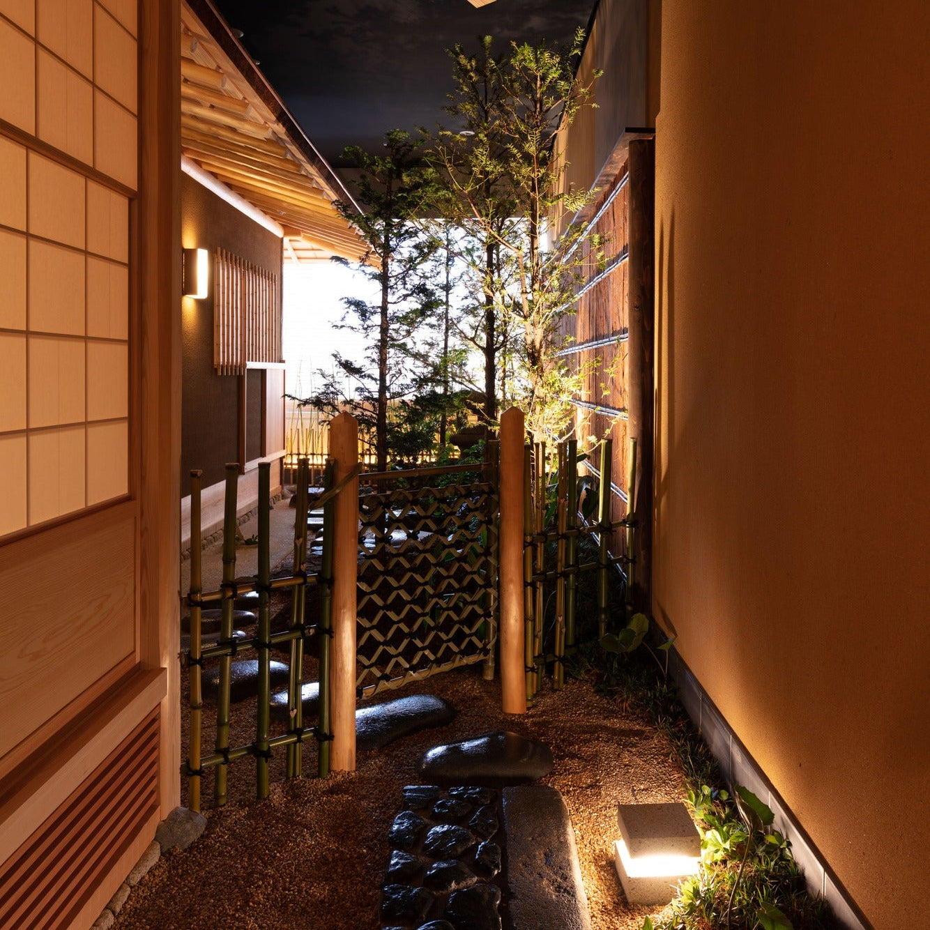 A narrow roji at night, stepping stones passing bamboo fences toward the chashitsu, lit low by a garden lantern.
