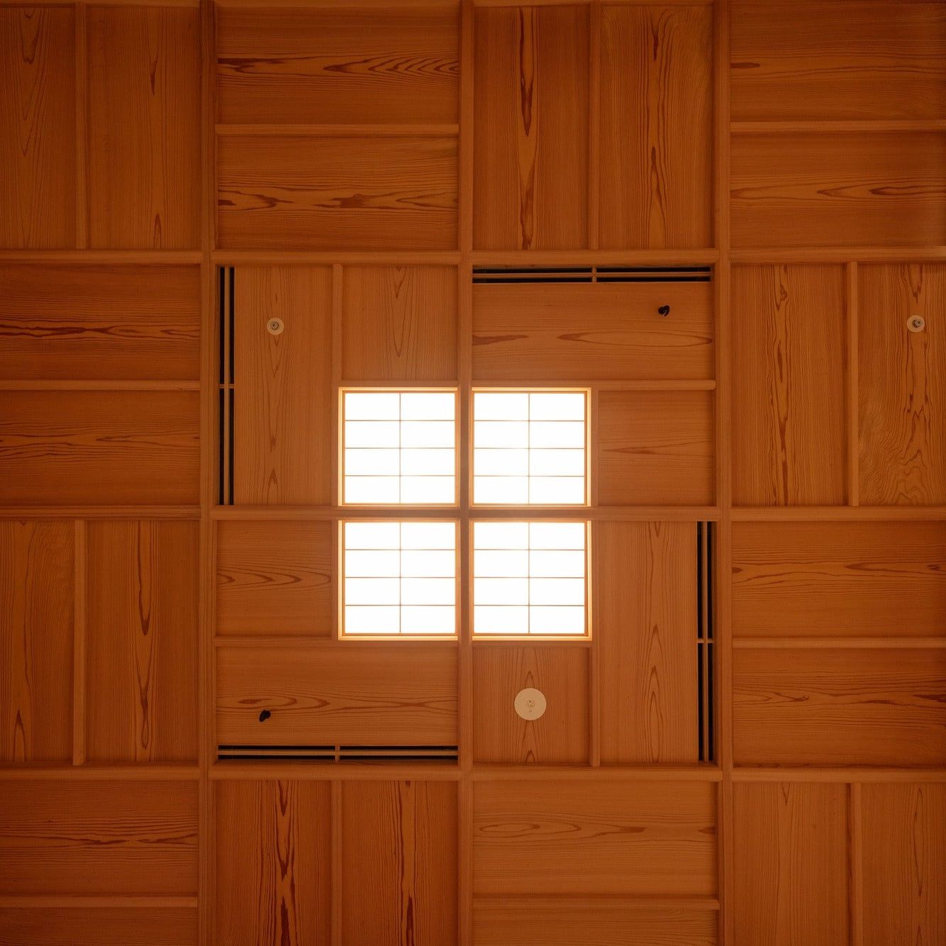 Grained timber boards wrap a wall and ceiling around a small square paper window that glows from within.