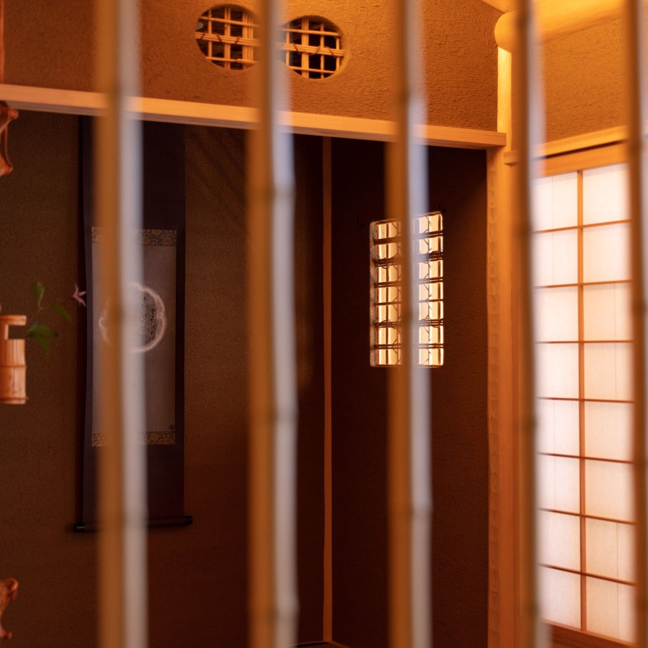 Through a lattice, a hanging scroll and a single flower in a bamboo vase rest in a dim alcove beside a round window.